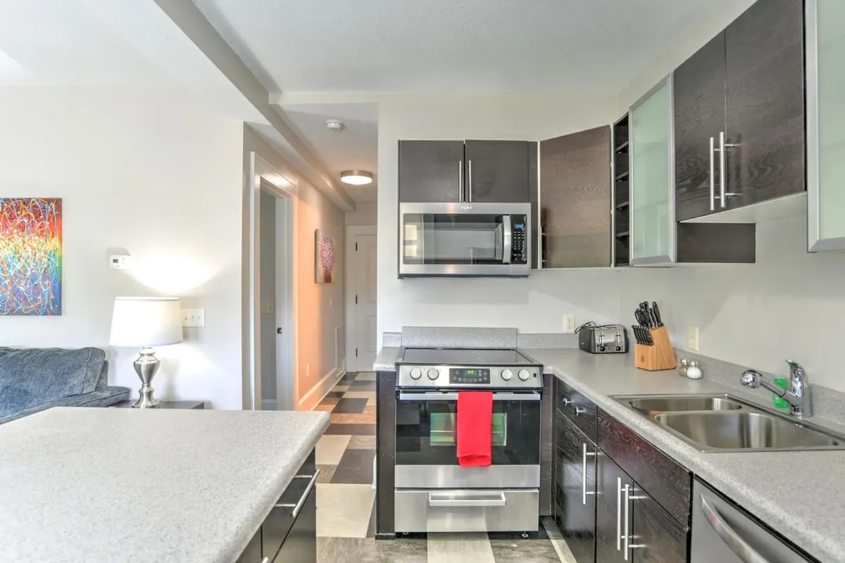 This image presents a well-lit, contemporary kitchen space featuring a central kitchen island, sleek stainless steel appliances, pops of color from wall art, and a bright red towel that adds energy to the scene. The layout is open and inviting, subtly hinting at gatherings, meal prep, and social culinary experiences.