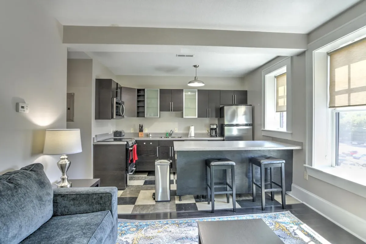 The image showcases a stylish, contemporary kitchen and living area with a central island and stools, sleek stainless steel appliances, warm lighting, and a cozy sofa—perfectly blending modern comforts with inviting charm.