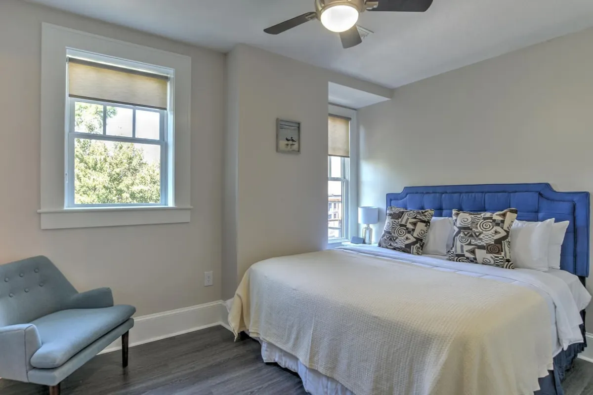 A tranquil bedroom scene featuring a neatly made bed with decorative pillows, an inviting armchair positioned near the window, and soft natural light streaming in. The room is styled with understated, tasteful decor and exudes a sense of quiet relaxation and warmth.